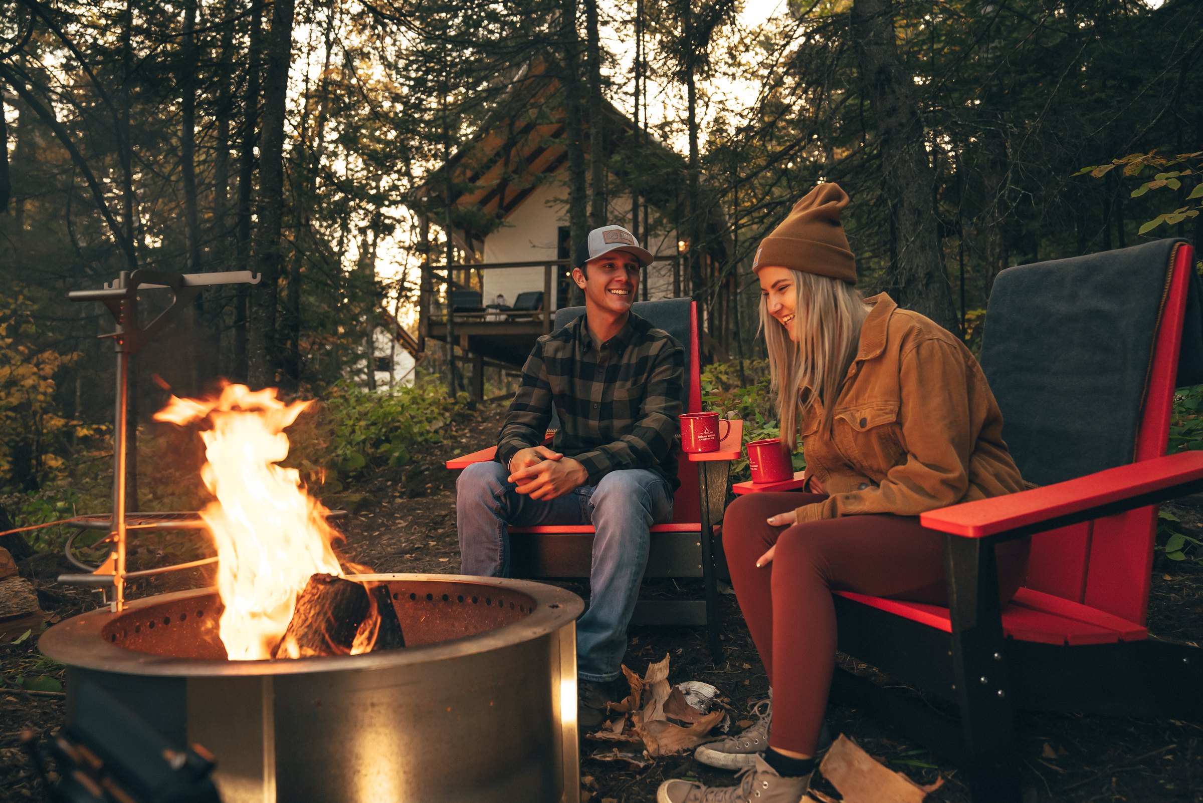 Two guests by a campfire in Adirondack chairs outside a cabin in the fall woods at North Shore Camping Co. in Beaver Bay, Minnesota