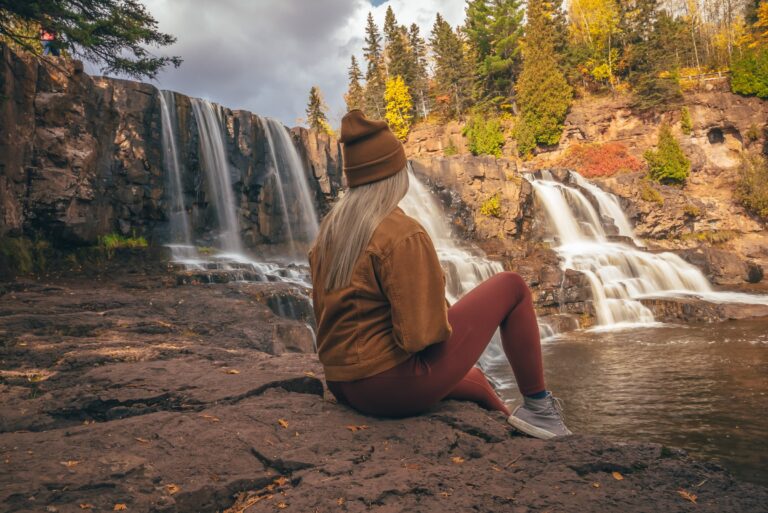 Visitor sitting on the rock ledge watching Gooseberry Falls flow over basalt with fall colors on the North Shore of Minnesota